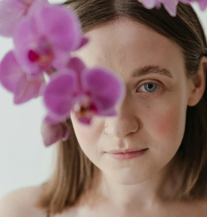 A close-up portrait of a young woman with fair skin and shoulder-length brown hair. She has a subtle nose ring and is gazing directly at the camera with a calm expression. The foreground features vibrant purple orchids, slightly blurred, framing her face and adding a soft, natural aesthetic. The background is bright and minimal, enhancing the delicate, serene mood of the photograph.
