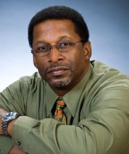 A professional headshot of a man with short black hair and a neatly trimmed goatee. He is wearing glasses, a green dress shirt, and a patterned tie. He has a thoughtful expression and is leaning slightly forward with his arms crossed. The background is a soft gradient of blue, creating a polished and professional atmosphere.