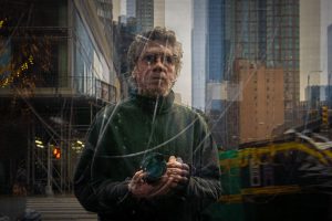 A man with curly hair, wearing a dark green jacket, holds a camera in front of his chest while standing in an urban environment. The image has a reflective overlay, capturing city buildings, scaffolding, and street elements, creating a layered effect. The background features tall skyscrapers and a blurred vehicle in motion, adding depth and movement to the composition.