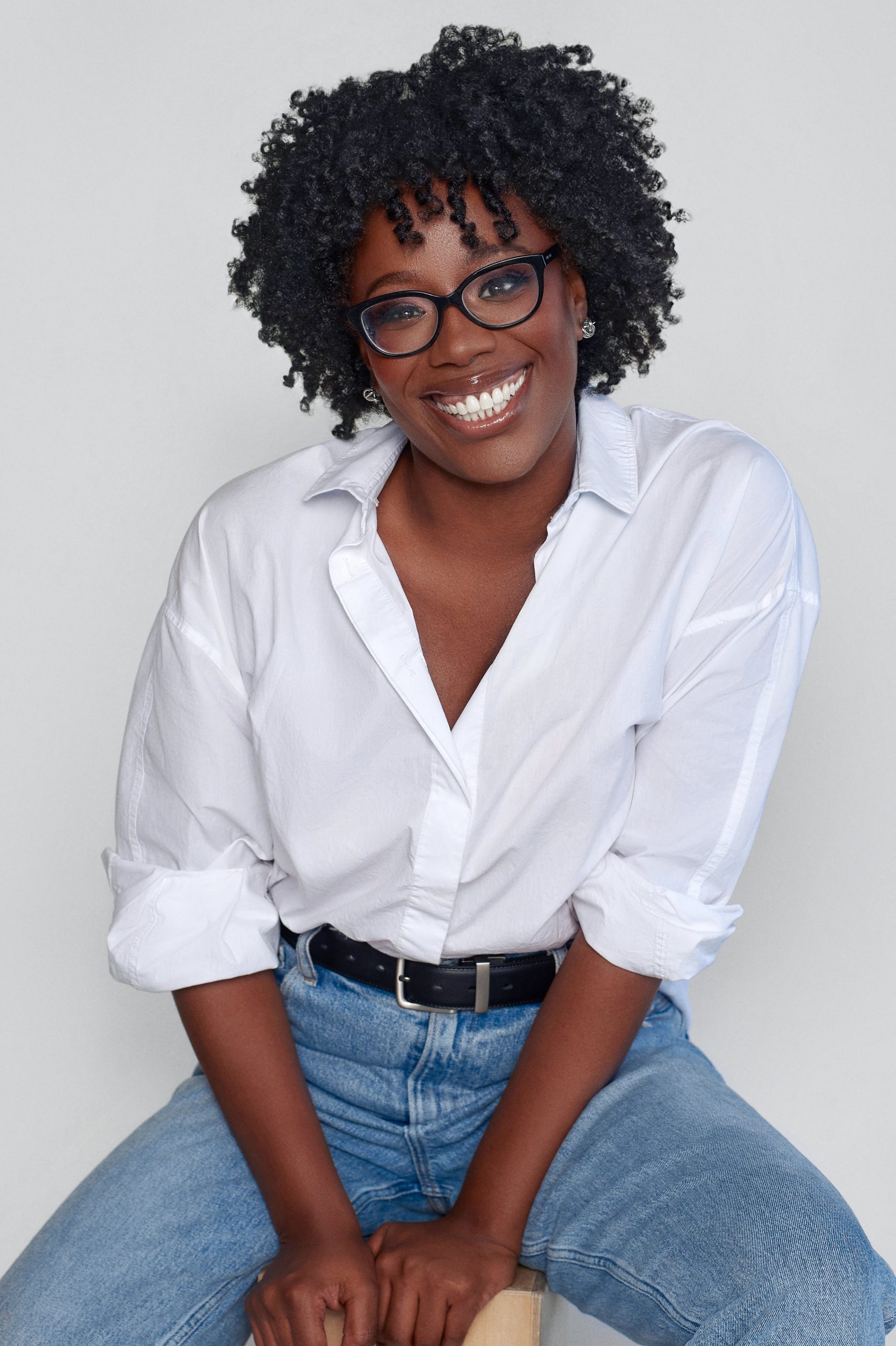 A photograph of a smiling Black woman with short, curly natural hair. She is wearing black-framed glasses, a white button-up shirt with rolled-up sleeves, and blue jeans with a black belt. She is sitting on a stool, leaning slightly forward with her hands resting on her thighs. The background is a plain light gray, and the lighting is soft and even.