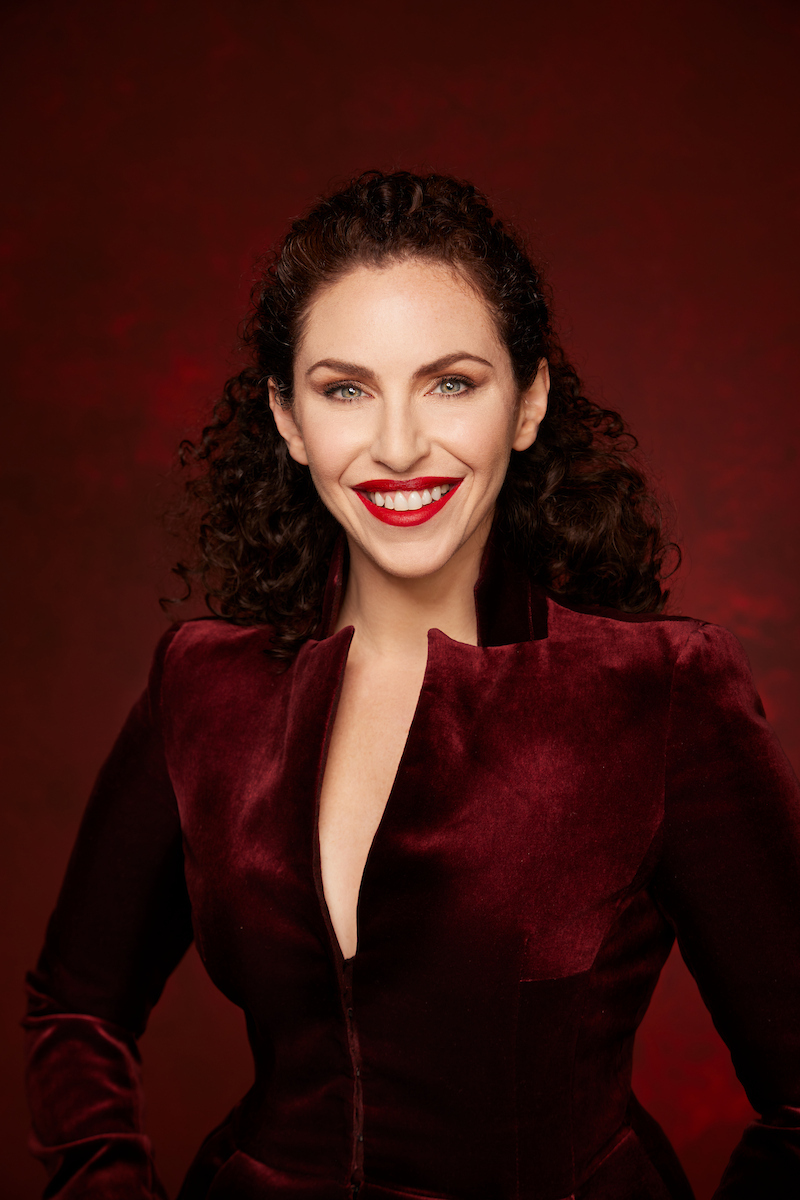 Portrait of Lindsay Adler, a woman, with fair skin and curly dark brown hair, smiling confidently. She is wearing a deep red velvet blazer with a plunging neckline. Her makeup features bold red lipstick and defined eyes. The background is a rich, dark red, complementing her attire. The lighting highlights her features, creating a dramatic and elegant effect.
