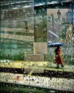 Image #9 by Margit Zsedely. A layered, collage-style photograph seen through a rain-covered window. Water droplets in sharp focus cover the glass, while blurred figures holding umbrellas walk outside. A person in a red coat with a blue umbrella appears in the lower right. Reflections of indoor elements—chairs, walls, and a large decorative object—overlay the rainy street scene, creating a dreamy, abstract composition of interior and exterior spaces merging together.